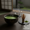 A traditional Japanese tea ceremony setting featuring a handmade ceramic bowl of frothy Grand Tea Ceremony Select matcha, a bamboo whisk (chasen), and a bamboo scoop (chashaku) on a minimalist wooden table, embodying the spirit of Wabi-cha